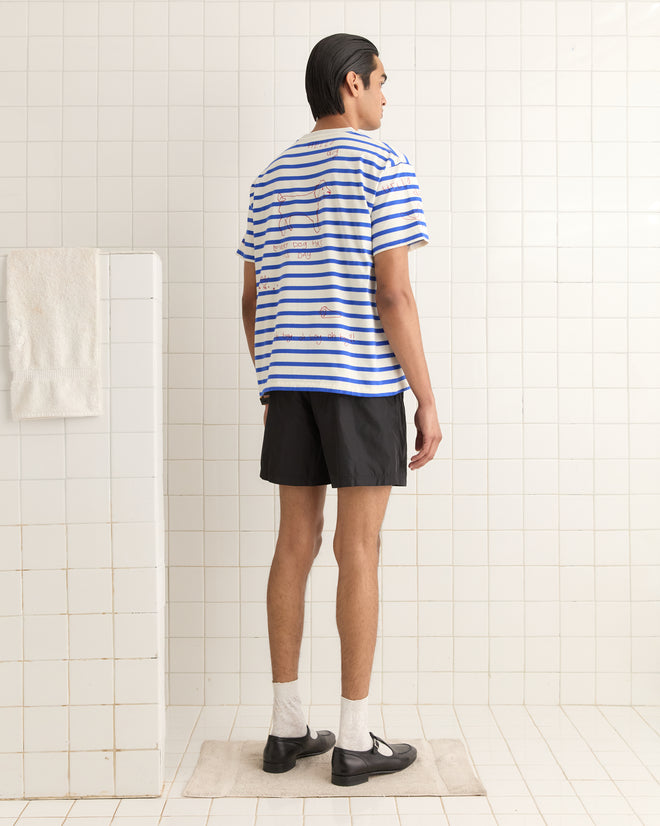 A person in a Margate Tee and black shorts stands on a bath mat in a tiled room, facing away.