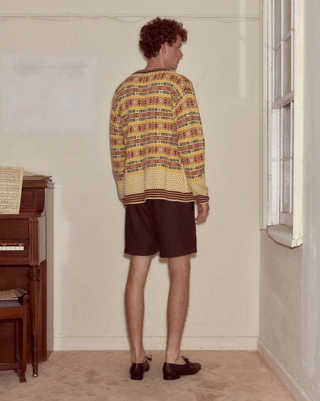 A man in the Marzipan Cardigan, featuring a Norwegian pattern and novelty silver ship buttons, stands in shorts facing the corner of a room with a piano to his left.