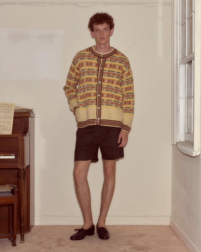 Wearing the Marzipan Cardigan with novelty silver ship buttons, brown shorts, and loafers, a person stands by a piano in a sunlit room.