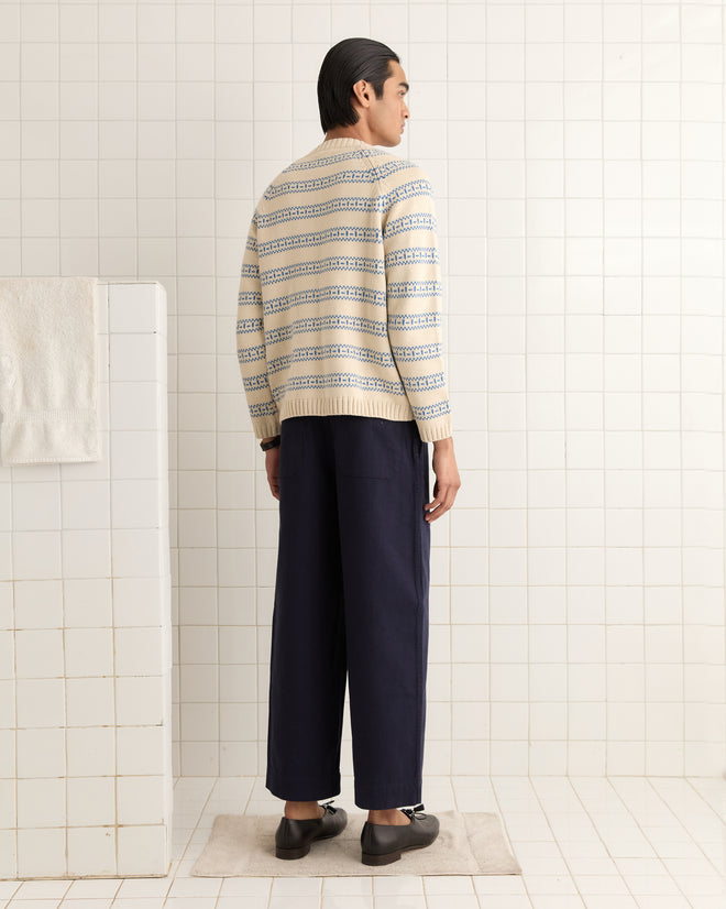 A person stands in a tiled room, wearing the Meandering Stripe Cardigan - Cream with dark pants and loafers, facing away from the camera.
