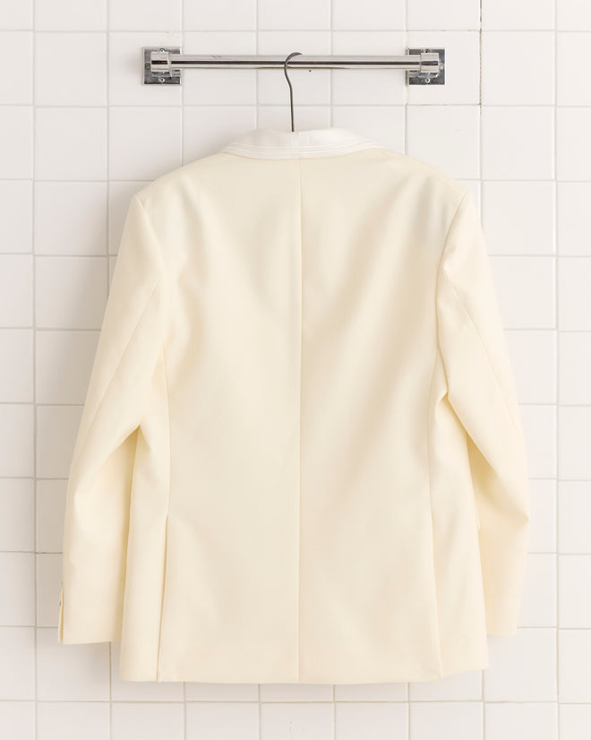 The Meringue Shawl Collar Tuxedo Jacket, crafted in ivory wool with a shawl collar, is displayed from the back on a hanger against a white tiled wall.
