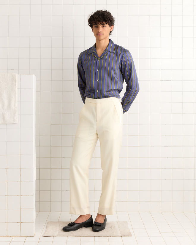 Wearing Meringue Smoking Trousers and a striped shirt, a person stands on a rug in a tiled room with hands behind their back.