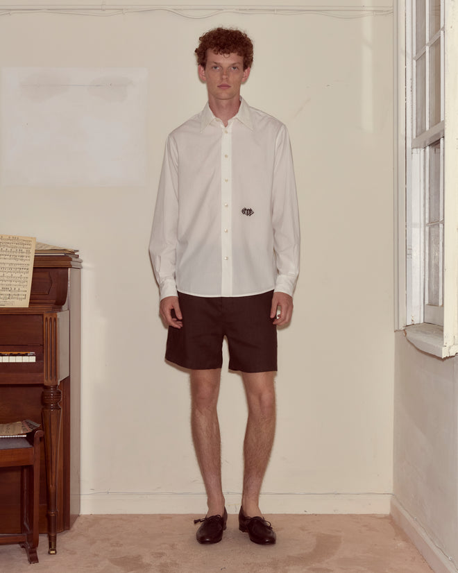 A man in a Monogrammed Poplin Shirt and black shorts stands by a piano in a sparsely decorated room.