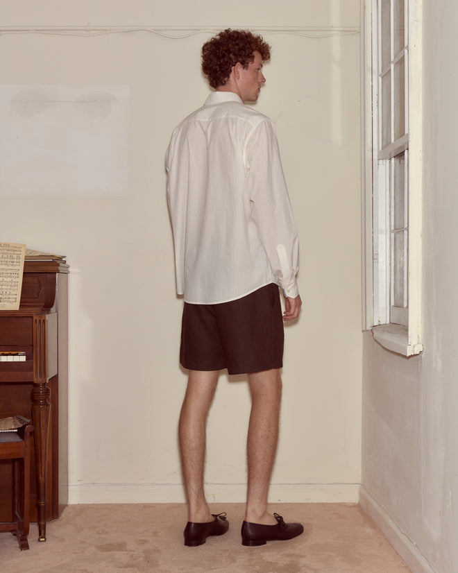 A man in a Monogrammed Poplin Shirt and black shorts stands facing a wall near a window and an upright piano.