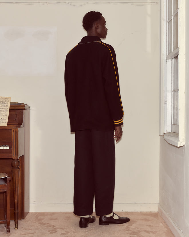 A person in a black Monogram Snap Reversible Jacket stands facing a cream wall near a piano and a window.