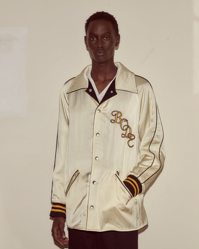 A person wearing the Monogram Snap Reversible Jacket—a shiny cream satin piece with black and gold striped cuffs and