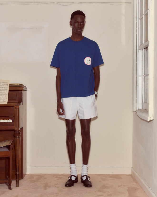A person wearing the Moon Tee, white shorts, socks, and black shoes stands by a piano in a bright room.
