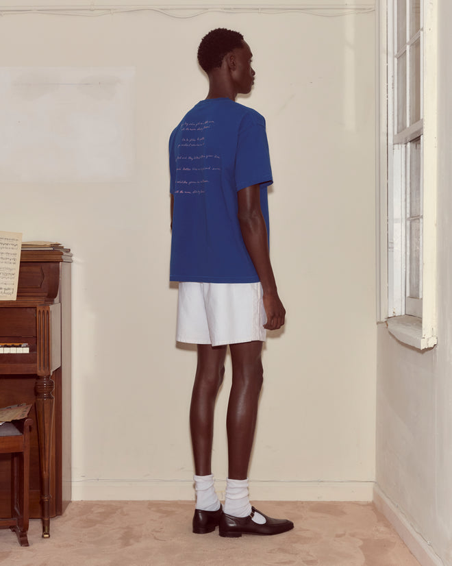 Wearing a Moon Tee, white shorts, and black shoes, a person stands in a corner near a window and piano.