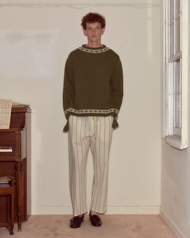 Indoors by a piano and window, a person wears the Moritz Sweater, crafted in green virgin wool, paired with white striped pants.