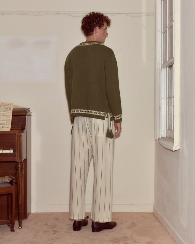 A man stands indoors facing a wall, wearing the Moritz Sweater in green Fair Isle and striped white pants, embodying timeless luxury knitwear.
