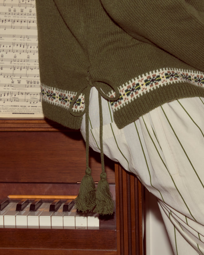 A person in striped pants and the Moritz Sweater sits at a piano with sheet music behind them, highlighting cozy luxury knitwear style.