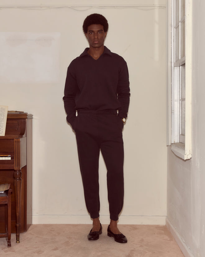A man in dark clothes, featuring Black Joggers, stands in a corner near a piano with sheet music, hands in pockets and looking forward.