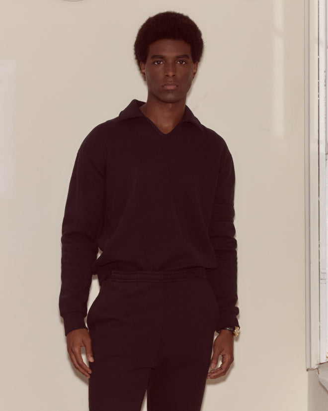 A man wearing the Crew Polo - Black stands indoors by a light wall near a window, looking at the camera.