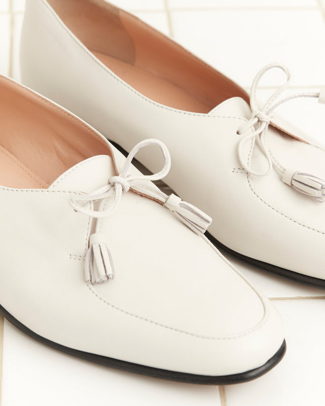 A pair of Tassel Shoes - White, featuring classic bow and tassel details, displayed on a light tiled surface.