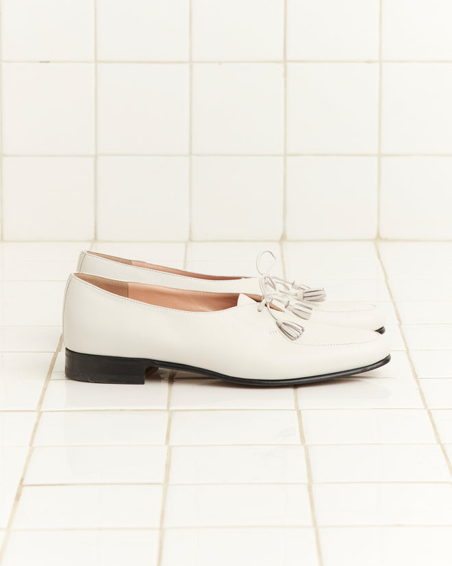 A pair of Tassel Shoes - White with black soles displayed on a white tiled floor and wall.