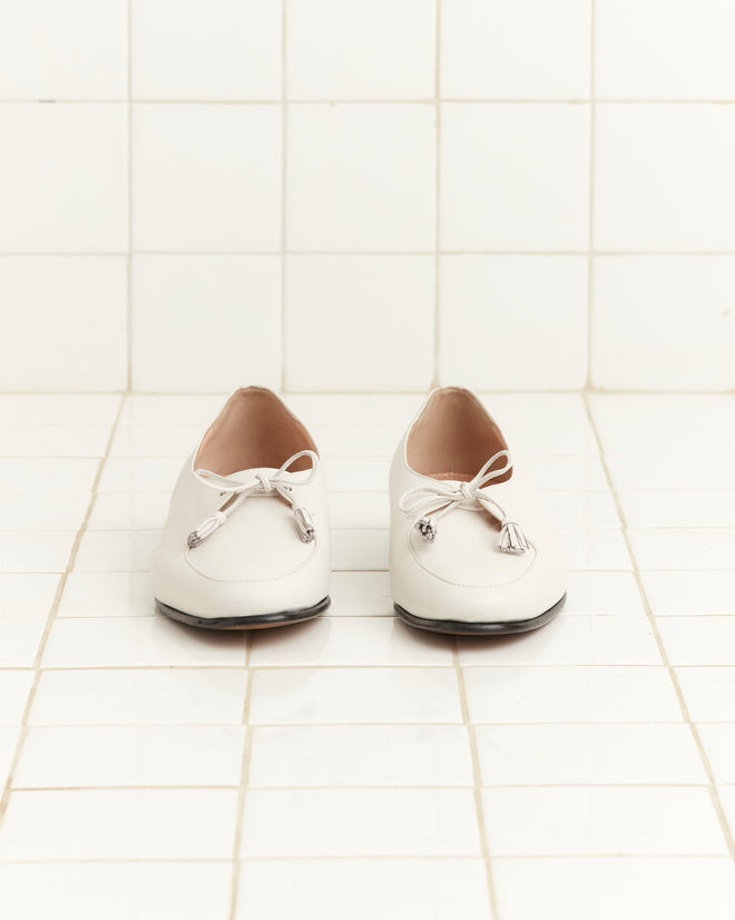 The Tassel Shoes - White are vintage leather loafers with tasseled ties, shown on a white tiled floor and wall.