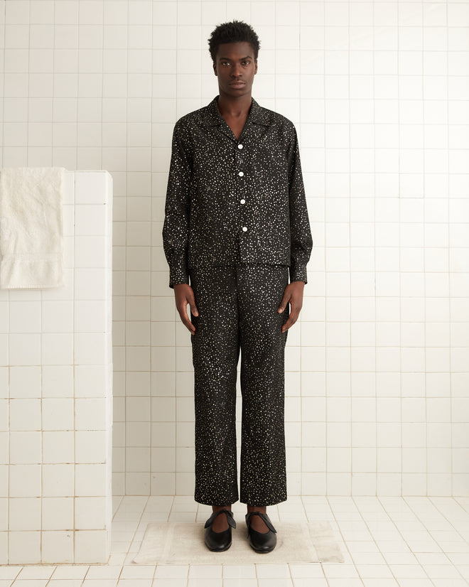 A man wearing a black Glitter Shirt stands in a white tiled bathroom with a towel hanging nearby.