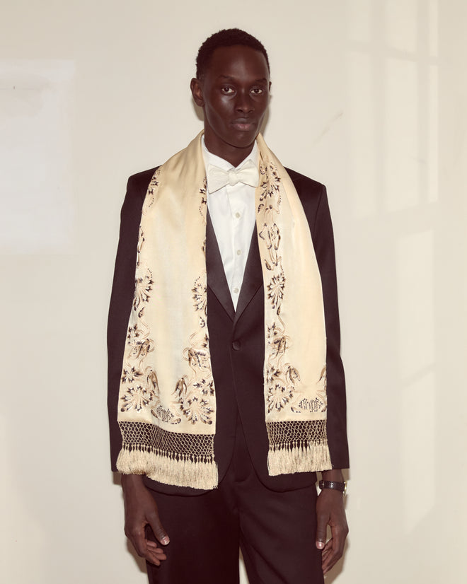 A person in a black suit and the Phantasm Fringe Scarf, featuring Victorian-inspired ivory silk embroidery, stands against a cream-colored wall.
