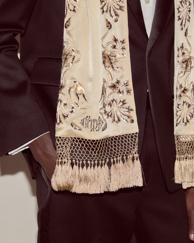 A person in a dark suit accessorizes with the Phantasm Fringe Scarf—an ivory silk piece featuring fringe and intricate floral patterns inspired by Victorian embroidery.