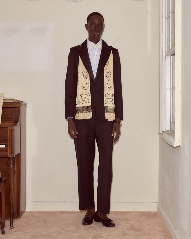 A man in a black suit with gold Victorian embroidery and a Phantasm Fringe Scarf stands indoors near a piano and a window.