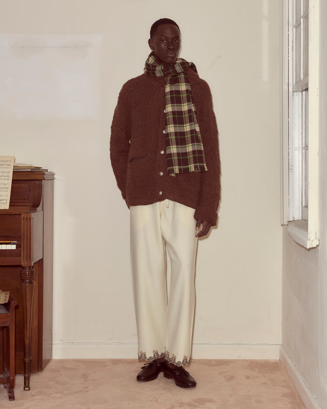 A person in a brown jacket, cream pants, Woodcrest Scarf, and black shoes stands indoors by a piano and window.