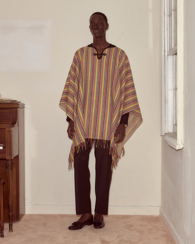 A person wearing the Lawrence Poncho with primary stripes and black pants stands indoors near a piano and a window.