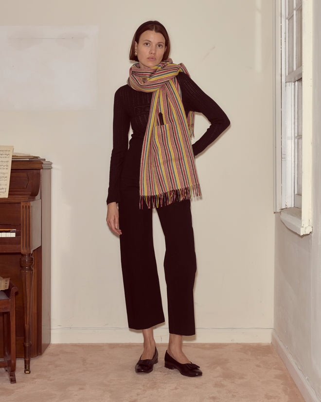 Woman in a black outfit and the Lawrence Shawl, featuring primary stripes, stands indoors near a piano, illuminated by natural light from the window.