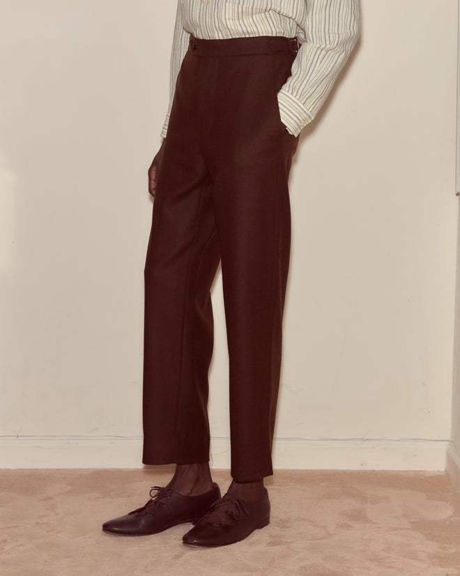 Wearing Bramble Side-Buckle Trousers in dark brown wool twill with a striped shirt and dark dress shoes on a beige carpet—an ensemble that captures classic suiting style.
