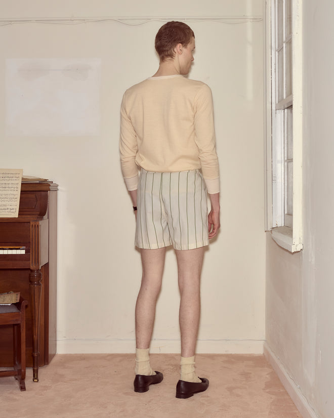 A person in a beige shirt and Celery Stripe Boxers, made from 100% cotton, stands facing a wall in a sparsely furnished room.