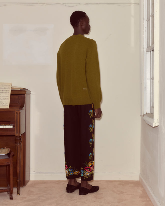 A person in a green sweater and Beaded Aquitaine Trousers stands facing a wall near a piano and window.