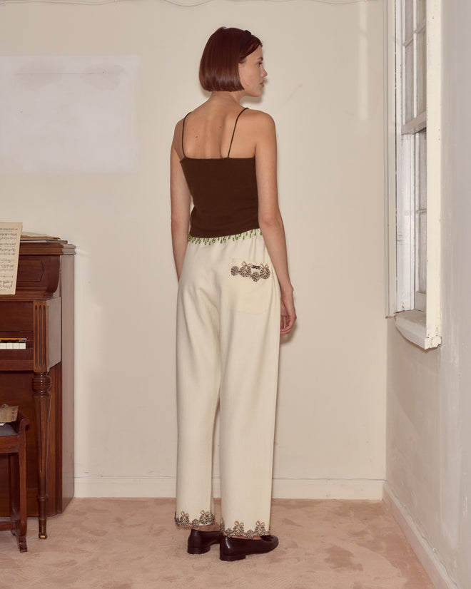 A woman stands facing away in a sparsely furnished room with a piano on the left, wearing a black top and Laurelist Pajama Pants - Cream.