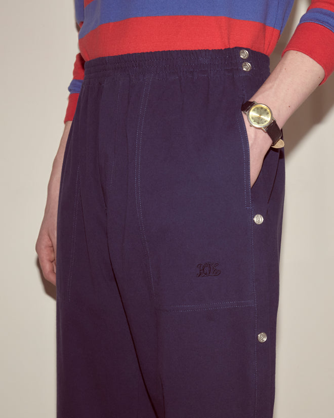 Model wears the Twill Tear-Away Track Pants - Navy, featuring side buttons, with a red-and-blue striped top, hand in pocket, and a watch.