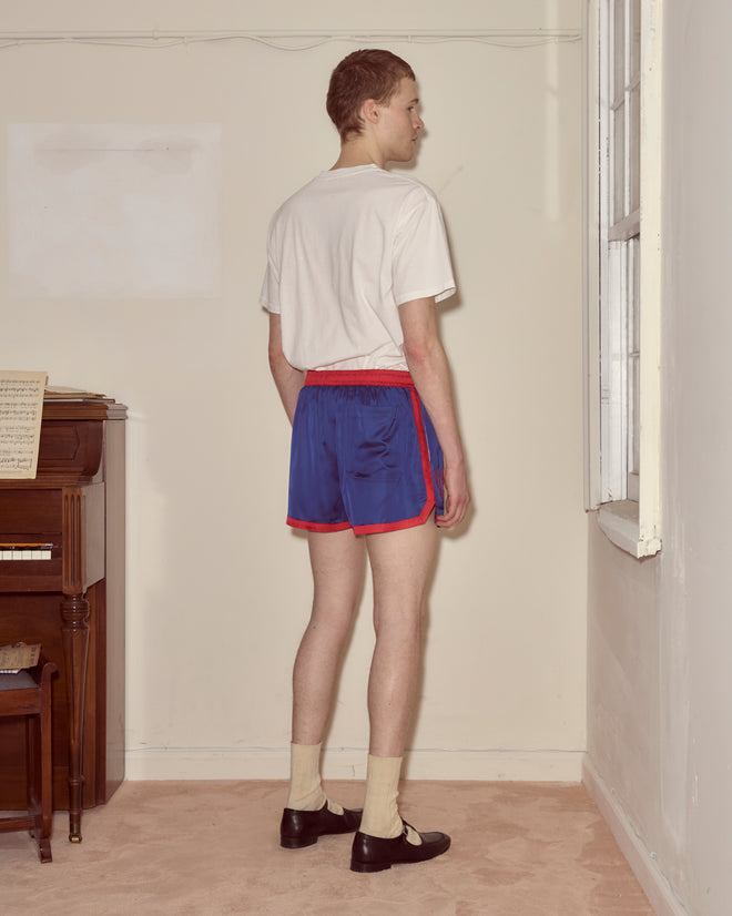 A person in a white shirt, Heart Deco Gym Shorts - Blue with a 1970s silhouette, and dress shoes stands near a window, facing away.