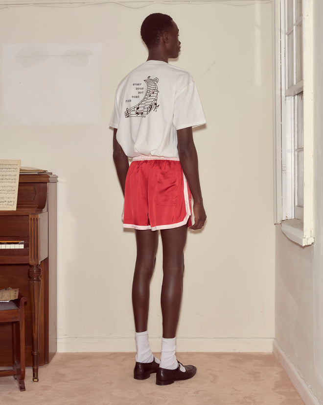 Indoors by a piano, a person wears Heart Deco Gym Shorts - Red, white socks, black shoes, and a graphic white Bode t-shirt—channeling a 1970s-inspired look.