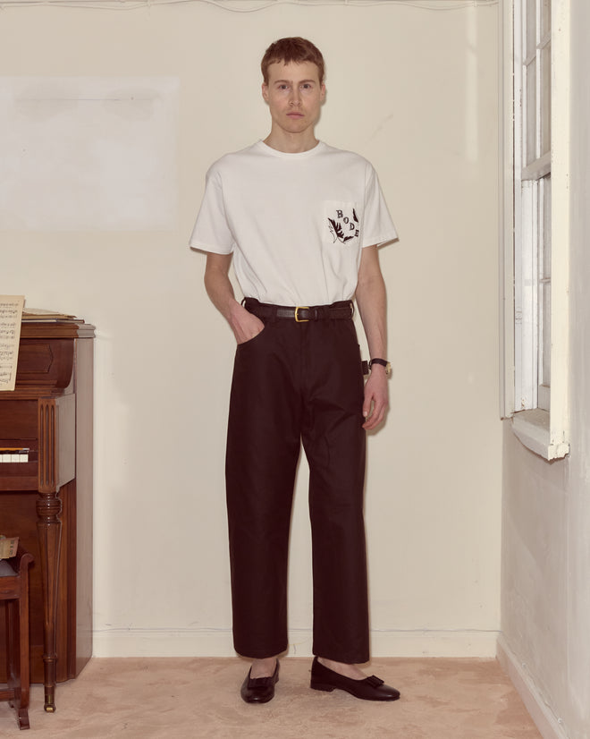 Man wearing a white t-shirt and Waxed Cotton Knolly Brook Trousers in brown stands indoors by a piano and window, looking at the camera.