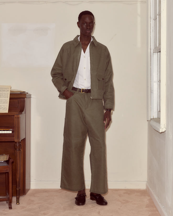 A man in an olive green suit and white shirt stands by a piano and window, his look completed with Waxed Cotton Knolly Brook Trousers in Light Green for a nod to timeless style.