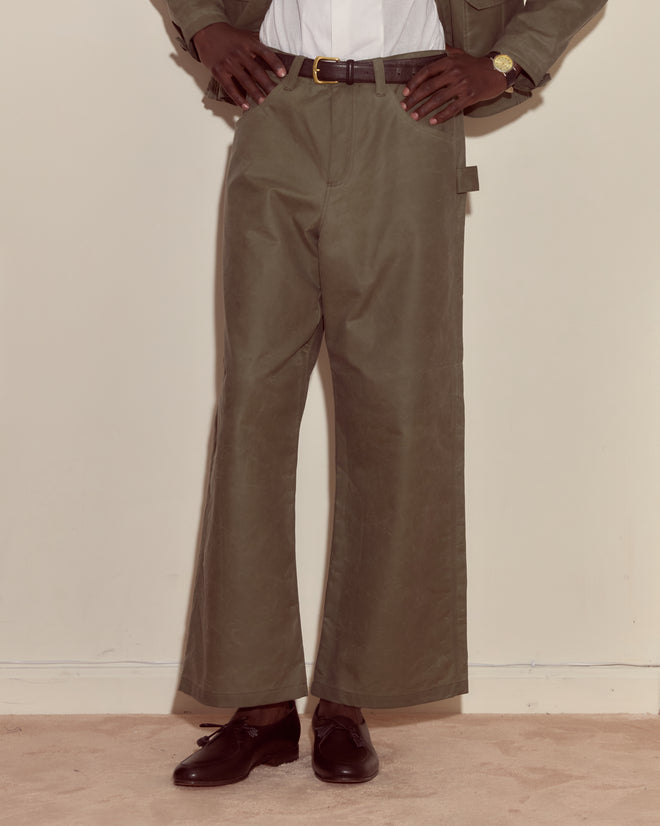 Person wearing Waxed Cotton Knolly Brook Trousers in light green, paired with a white shirt, brown belt, and brown shoes, hands resting on hips.