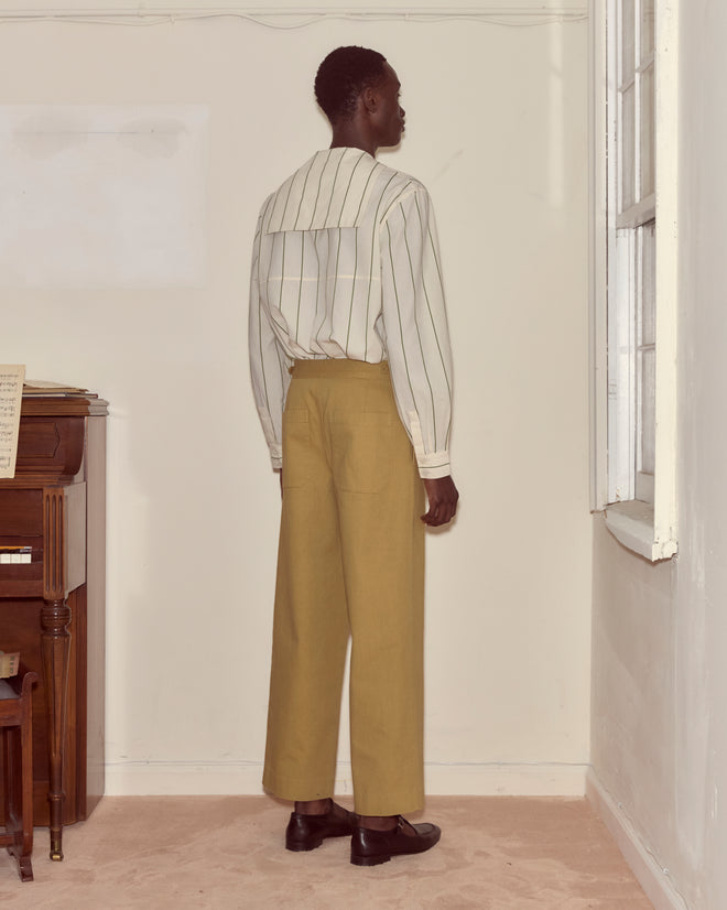 Person standing indoors facing away, wearing a striped shirt and Wide Leg Snap Trousers - Khaki, next to a piano.
