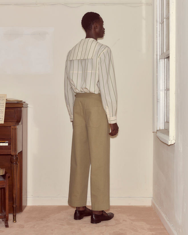 A person stands indoors facing a wall, wearing a striped shirt and the Wide Leg Snap Trousers - Khaki, featuring vintage-inspired double front pleating.
