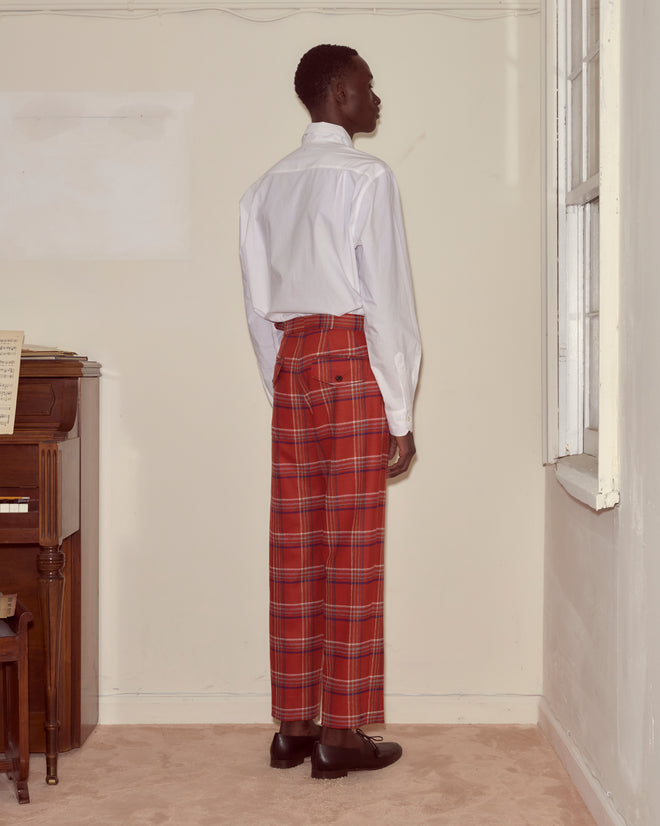 Wearing a white shirt and Gable Plaid Trousers, a person stands facing the corner in a sparsely furnished room.