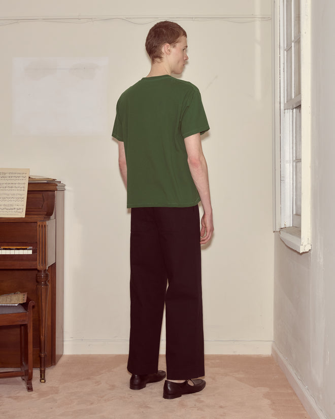 Someone wearing a Pirate Ship Tee and black pants stands facing a corner near a window and a piano.