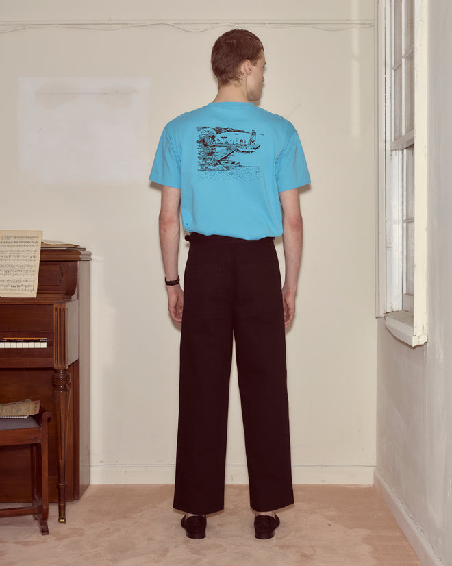 Person in a blue Laketown Tee and black pants stands facing a wall in a room with a piano and window, evoking memories of Camp Onibar in the Poconos.