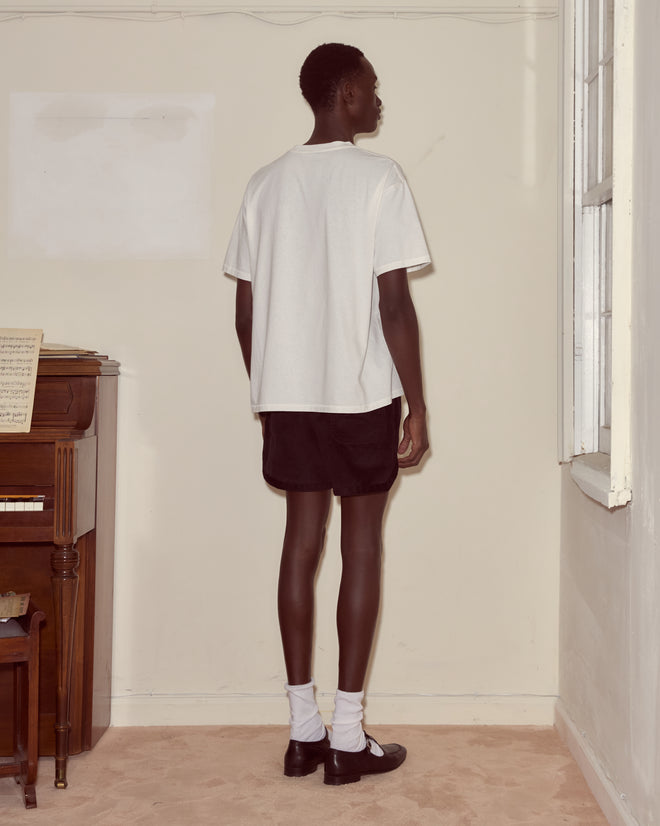 A person wearing the Chapeau Tee in white with black shorts stands facing a wall beside a window and a piano.