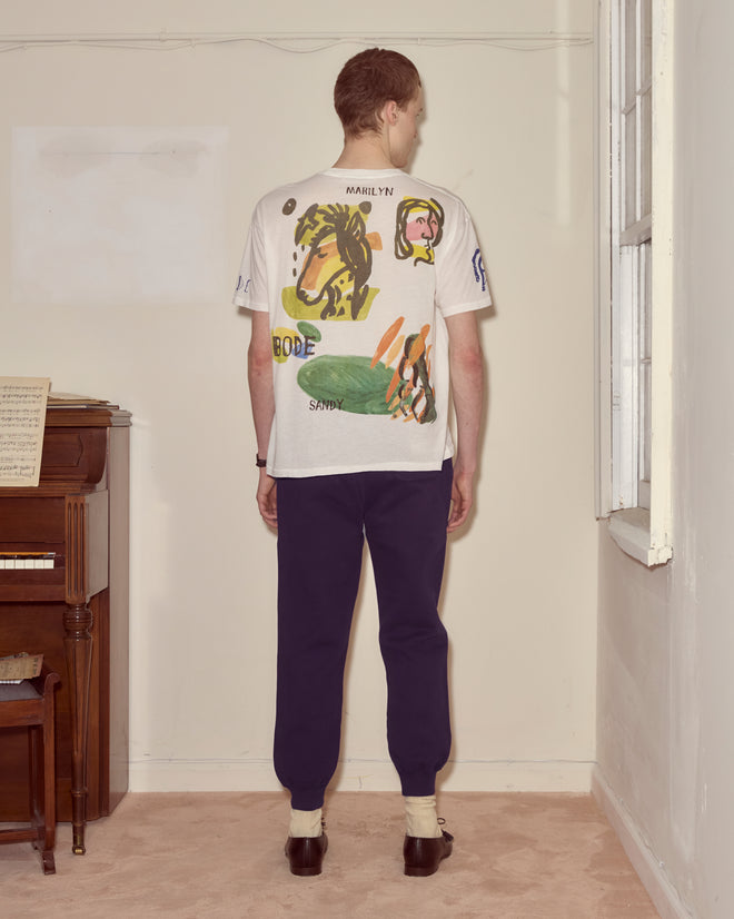 A person stands indoors, facing away, wearing the Collage Tee with dark pants and socks with sandals—an ensemble subtly echoing modernist art’s flair for abstract colorblocking.
