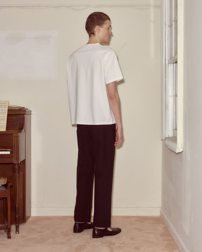 A person in a white Beaded Leaf Pocket Tee and black pants stands facing a wall near a window and a piano.