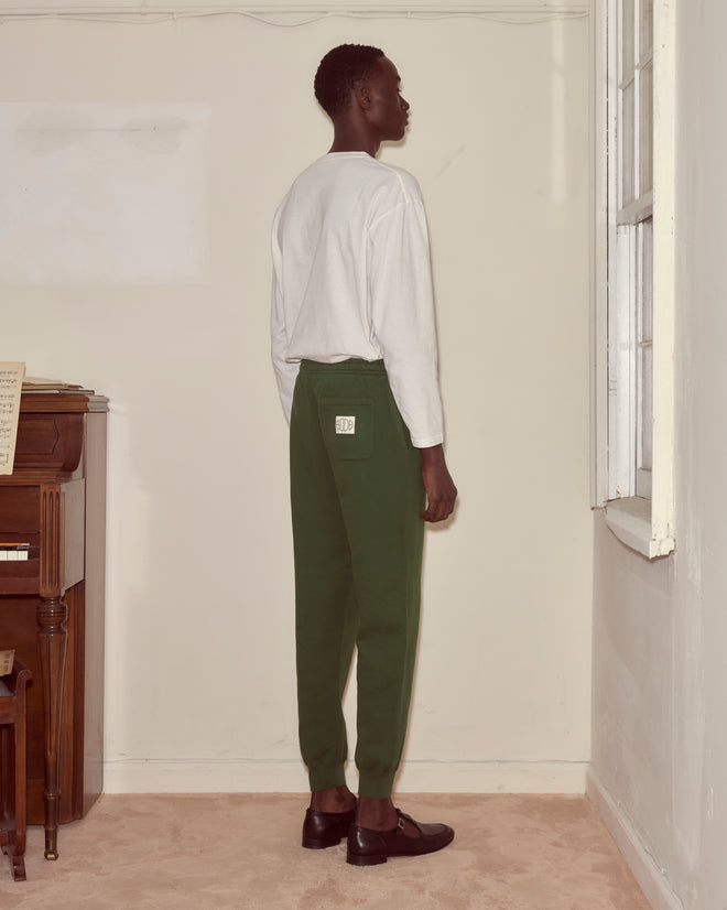 A person in a white shirt and Joggers - Dark Green stands facing a cream-colored wall near a window and piano, the look completed with a Bode woven label detail.