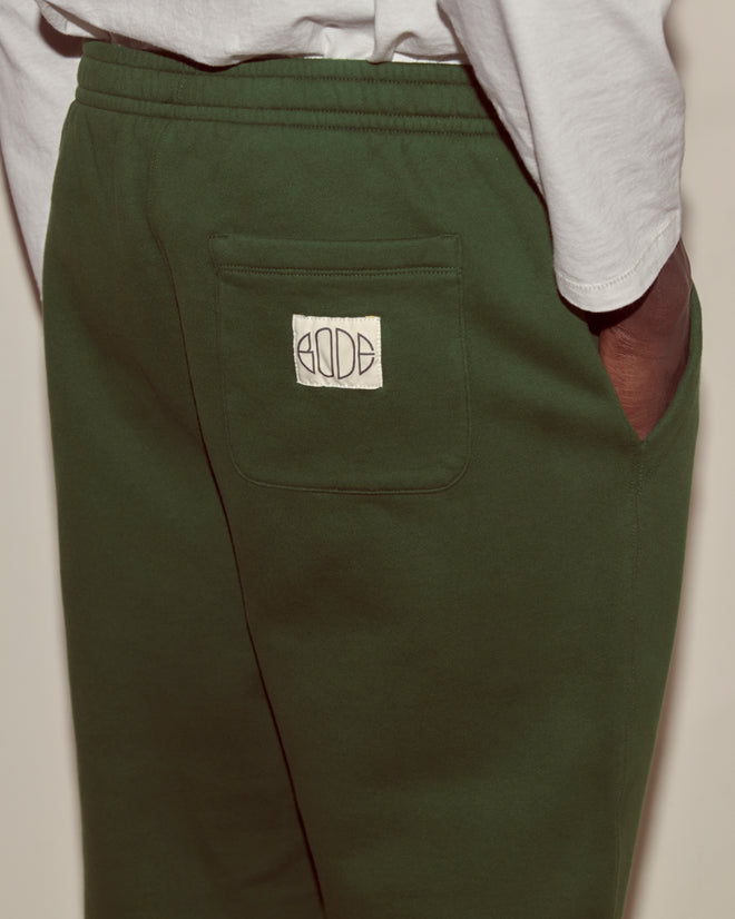 A person wearing Joggers - Dark Green midweight fleece cotton pants featuring a white Bode woven label on the back pocket.