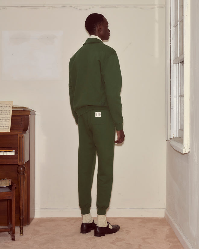 A person wearing a dark green Crew Polo stands facing a beige wall near a window and an upright piano with sheet music.