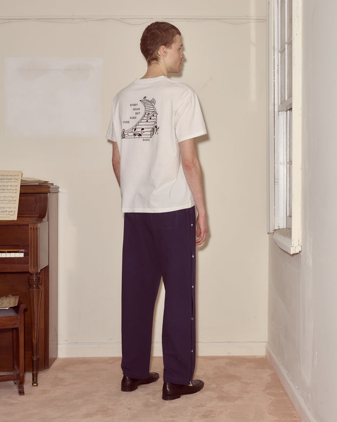 A person stands indoors facing a beige wall, wearing the Composer Tee and dark pants.
