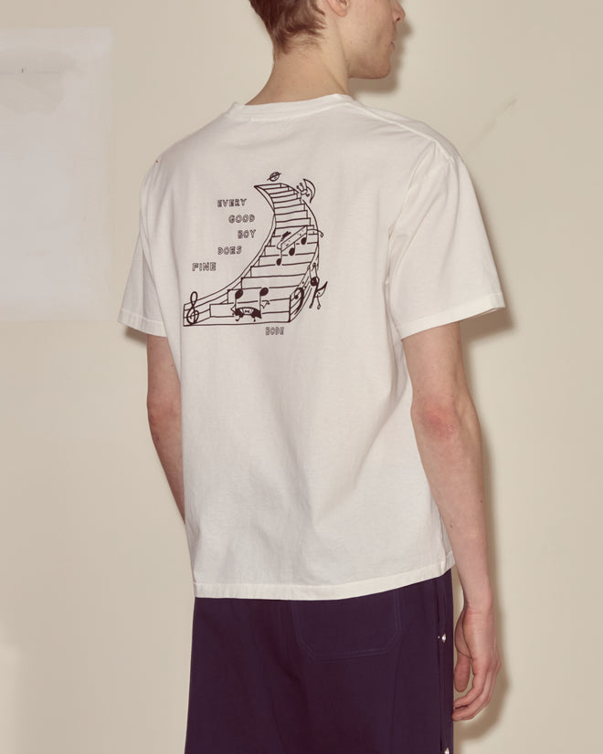 A person wearing the Composer Tee, a white embroidered shirt with a musical staff and vintage music notes graphic on the back.